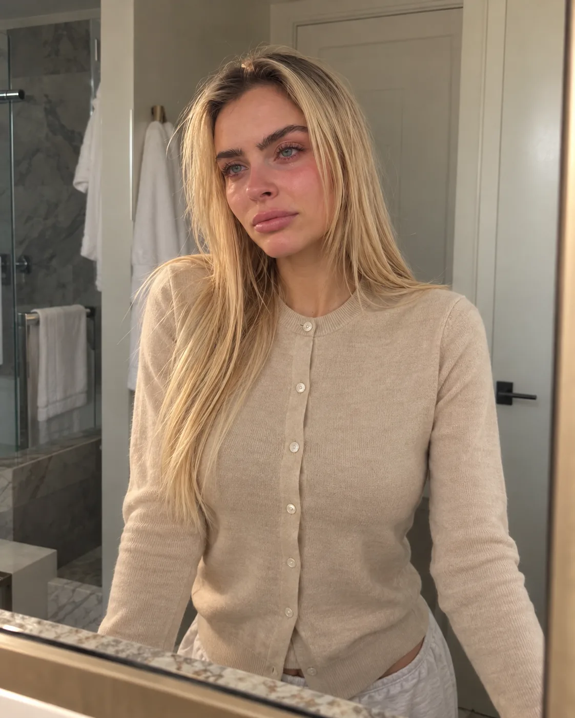 Katie in a beige cardigan buttoned to the neck, standing at a bathroom mirror